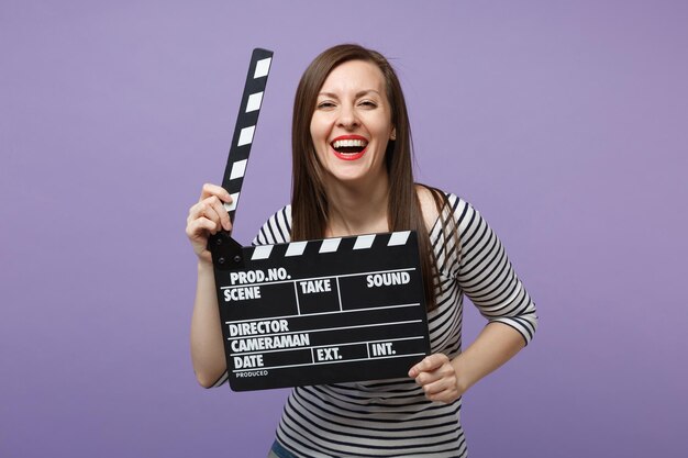 Garota jovem divertida em roupas listradas casuais posando isolado no retrato de estúdio de fundo roxo violeta. Conceito de estilo de vida de emoções sinceras de pessoas. Mock up espaço de cópia. Segure a claquete para fazer filme