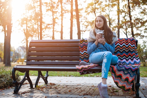 Garota jovem e atraente, sentado em um banco no parque outono e tomando café