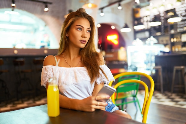 Garota jovem e bonita com um sorriso encantador, sentado em um café, bebendo limonada e usando um telefone