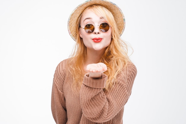 Garota jovem elegante atraente hipster em óculos escuros e chapéu de palha manda beijo no ar para a câmera. mulher divertida e feliz com lábios vermelhos na parede cinza com espaço de cópia