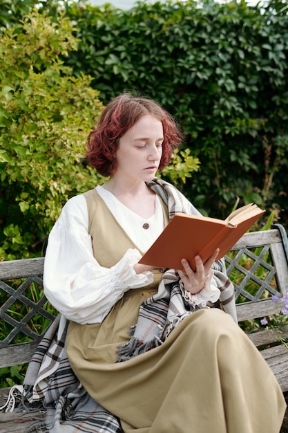 Garota jovem em casualwear lendo livro enquanto está sentado no banco no jardim