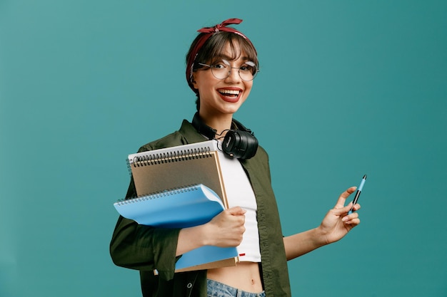 Foto garota jovem estudante alegre usando óculos de bandana e fones de ouvido no pescoço em pé na vista de perfil, olhando para a câmera segurando blocos de notas grandes e caneta isolada em fundo azul