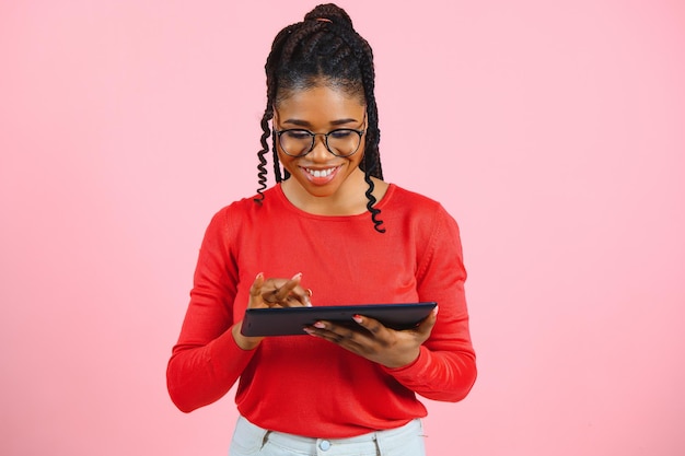 Garota jovem estudante americana com cabelo africano encaracolado segurando o tablet digital e sorrindo em pé sobre fundo rosa isolado com espaço de cópia para logotipo de texto ou publicidade