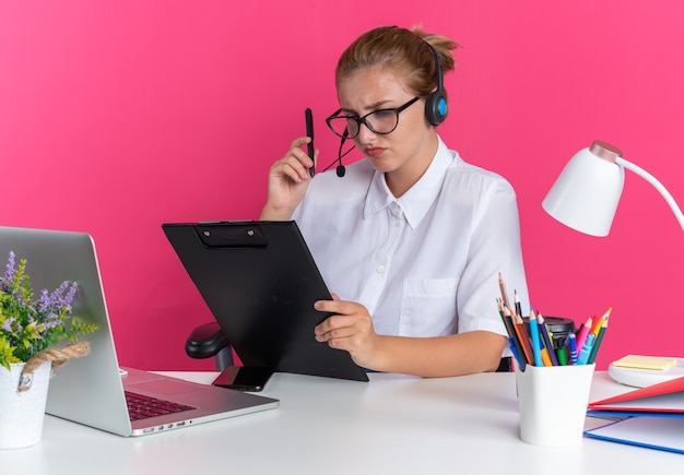 Garota jovem loira confusa em call center usando fone de ouvido e óculos, sentada na mesa com ferramentas de trabalho, segurando uma caneta e uma prancheta, olhando para a área de transferência isolada na parede rosa
