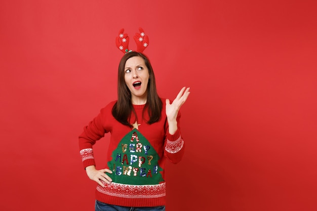 Garota jovem Santa indignada em diversão decorativos chifres de veado na cabeça, olhando para cima, levantando a mão isolada no fundo da parede vermelha brilhante. Conceito de festa de feriado de celebração de ano novo feliz 2019. Simule o espaço da cópia.