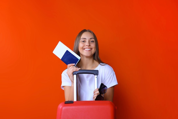 Garota jovem turista em roupas casuais de verão, com óculos de sol, mala vermelha, passaporte isolado em fundo vermelho.