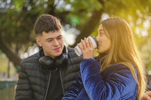 Garota latina bebe café em um copo descartável ao lado de um garoto caucasiano no banco de um parque público
