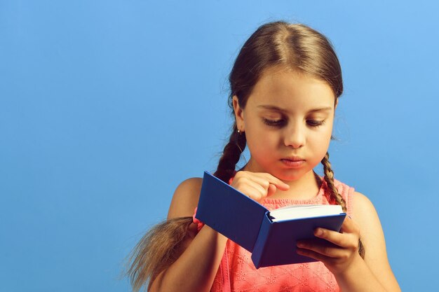 Garota lê livro azul garota da escola com rosto concentrado isolado em fundo azul de volta ao conceito de escola e educação garoto de vestido rosa com tranças e notebook