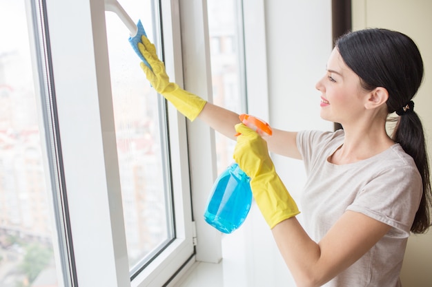 Garota legal e agradável fica na frente da janela e limpá-lo com pano e spray líquido azul. Menina usa luvas amarelas.