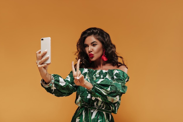 Garota legal e encantadora com cabelo escuro encaracolado em vestido de verão impresso branco e verde mostrando sinal de paz e faz foto em pano de fundo laranja