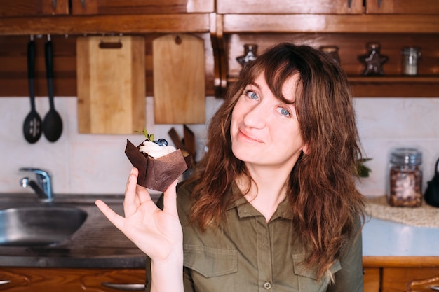 Garota legal sorri e detém cupcake e bata na cozinha de casa.