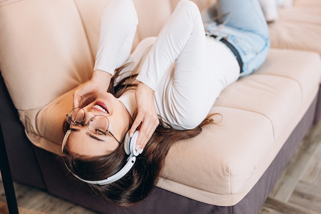 Garota legal usando fones de ouvido e ouvindo música em casa deitada no sofá
