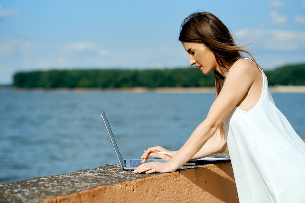 Garota linda e séria em um vestido branco com um tablet, caneta, telefone portátil no banco do réus. Foto de alta qualidade