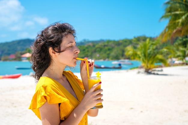 Garota linda em um vestido amarelo bebe manga fresca na praia de uma ilha paradisíaca. Férias perfeitas.