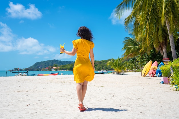 Garota linda em um vestido amarelo bebe manga fresca na praia de uma ilha paradisíaca. Férias perfeitas.