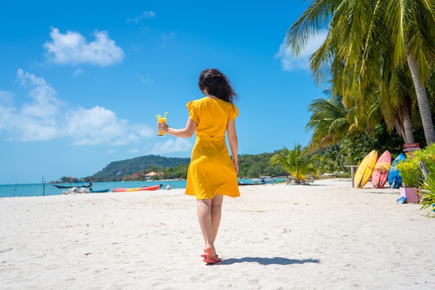Garota linda em um vestido amarelo bebe manga fresca na praia de uma ilha paradisíaca. Férias perfeitas.