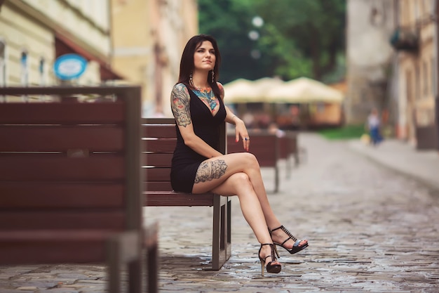 Garota linda em um vestido preto, sentado em um banco, coberto de tatuagens