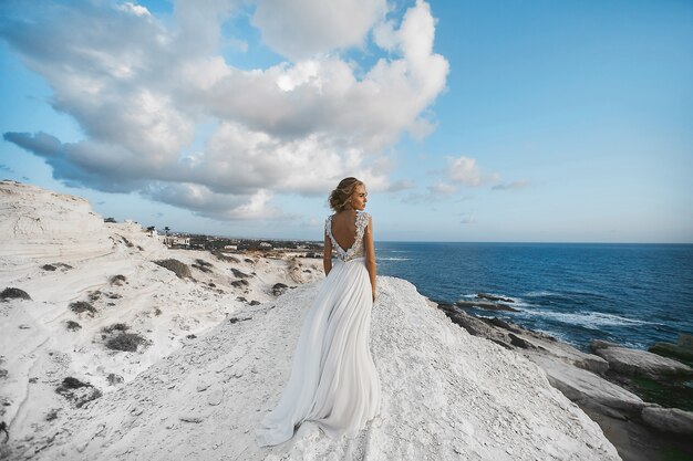 Garota linda jovem modelo loira, vestido branco, fica de pé sobre a pedra branca e olha para o mar