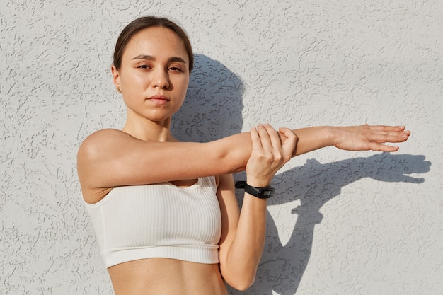 Garota linda modelo fitness vestindo roupas esportivas posando contra uma parede cinza ao ar livre, se aquecendo antes do treino