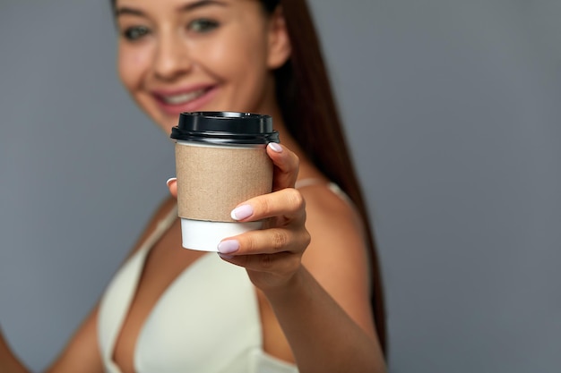 Garota linda mulher gosta e oferece uma bebida quente, chá ou café em um copo natural descartável em um fundo cinza claro conceito começar o dia com uma xícara de café copiar espaço cinza de fundo