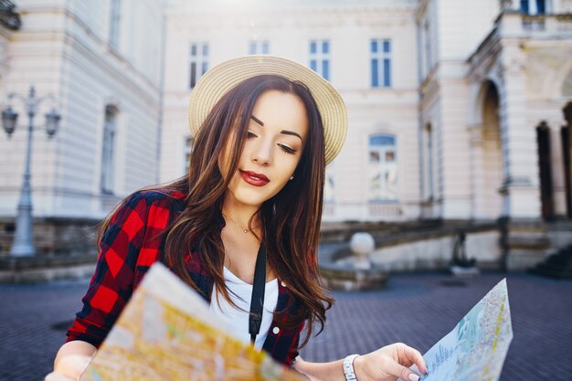 Garota linda turista com cabelo castanho, usando chapéu e camisa vermelha, segurando o mapa no antigo fundo da cidade europeia e sorrindo, viajando.