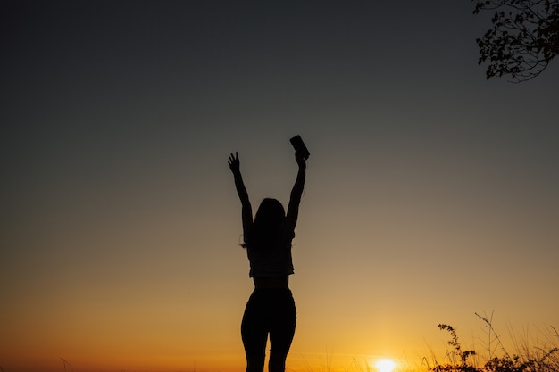 Garota livre em fones de ouvido, ouvir música e dançar com as mãos ao alto ao pôr do sol no campo.