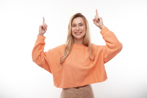 Garota loira alegre em sorriso casual laranja aponta o dedo para o espaço de cópia vazio para texto ou produto isolado no banner de publicidade de fundo branco