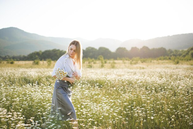 Garota loira atraente pegar flor de camomila andar no campo sobre a natureza verde ao ar livre Linda mulher segurando o buquê de camomila no Prado sobre a luz do sol fora da primavera