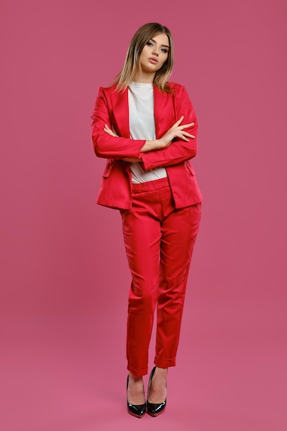 Garota loira de terninho vermelho, blusa branca, salto alto preto, posando em pé contra o fundo rosa do estúdio