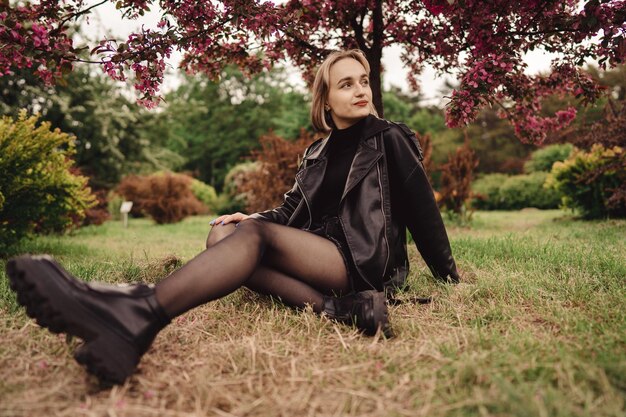 Foto garota loira e bonita de preto senta-se no chão no parque e relaxa sob a árvore de folhagem rosa