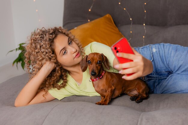 Garota loira encantadora com um cachorro bassê e tira uma selfie em um smartphone em uma caixa vermelha