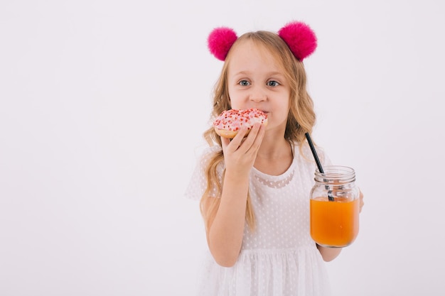 Garota loira engraçada comendo um donut e bebendo suco de frutas em um fundo branco