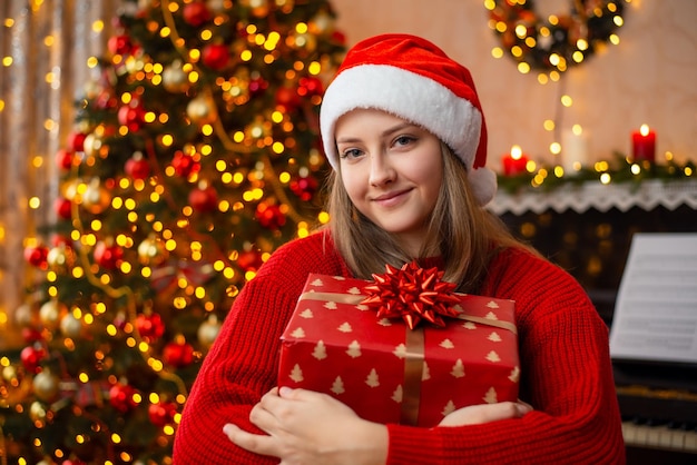 Garota loira feliz abraça grande caixa de presente vermelha Árvore de Natal decorada e luzes de guirlanda no fundo atmosfera aconchegante em casa feliz Natal e feliz ano novo conceito