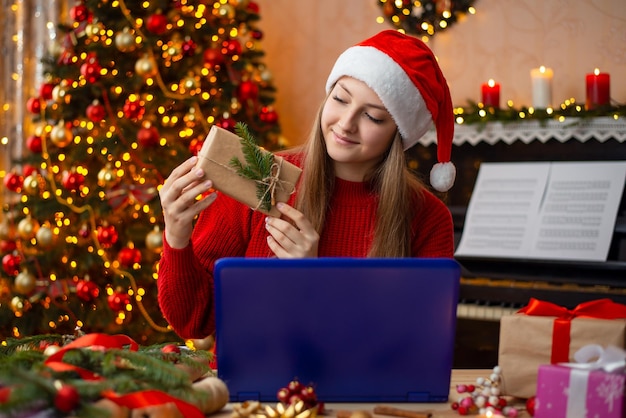 Garota loira feliz no chapéu de Papai Noel conversando com um amigo via internet no laptop e mostrando a caixa de presente que ela fez feliz Natal e Ano Novo conceito de amizade online de comunicação