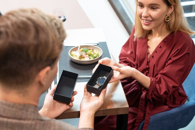 Foto garota loira feliz passando um relógio de pulso em uma pequena caixa de presente para o namorado e olhando para ele enquanto os dois estão sentados à mesa em um restaurante