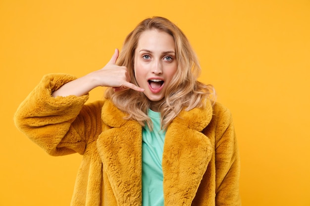 Garota loira jovem atraente com casaco de pele amarelo posando isolado no retrato de estúdio de fundo laranja. Conceito de estilo de vida de pessoas. Mock up espaço de cópia. Fazendo gesto de telefone como diz me ligue de volta.