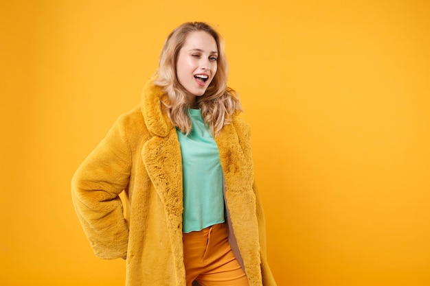 Garota loira jovem impressionante engraçada com casaco de pele amarelo posando isolado no retrato de estúdio de fundo laranja. Conceito de estilo de vida de emoções sinceras de pessoas. Mock up espaço de cópia. Câmera olhando piscando.