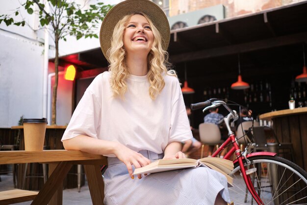 Garota loira muito alegre de chapéu alegremente sentada com livro e bicicleta no pátio do café da cidade