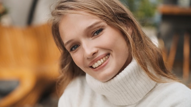 Garota loira muito sorridente vestida com um suéter branco aconchegante parecendo feliz posando na câmera ao ar livre