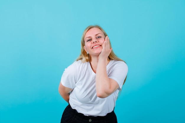 Garota loira sorridente está posando para a câmera, colocando a mão na bochecha no fundo azul