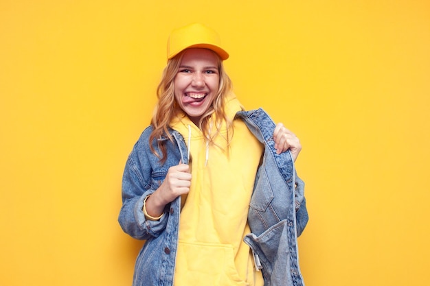 Garota maluca em jaqueta jeans e boné posando em fundo amarelo isolado garota hipster engraçada sorrindo