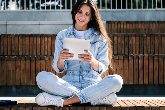 Garota milenar em roupas jeans se senta no banco do parque, segurando um tablet inteligente nas mãos. mulher usa tablet