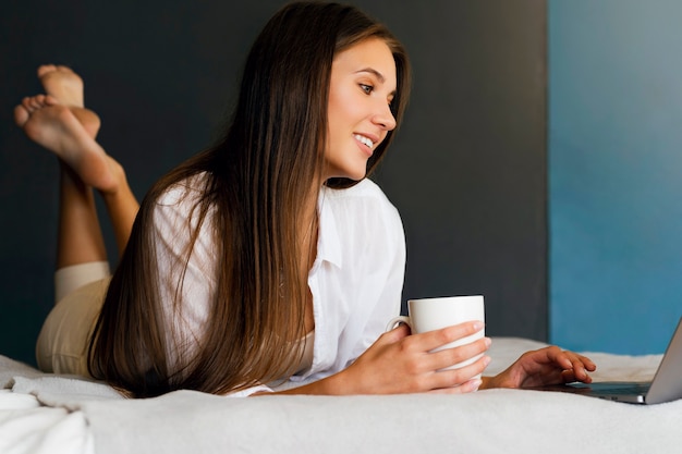 Foto garota milenar está descansando na cama em uma camisa branca, segurando uma xícara de café na mão.