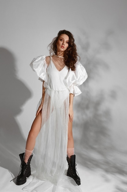 Garota modelo atraente em vestido branco com saia translúcida e botas de couro preto na moda no qu...