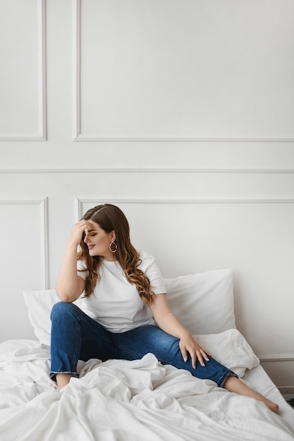 Foto garota modelo plus size em jeans e camiseta branca em branco, sentada na cama. moda xxl. beleza unideal