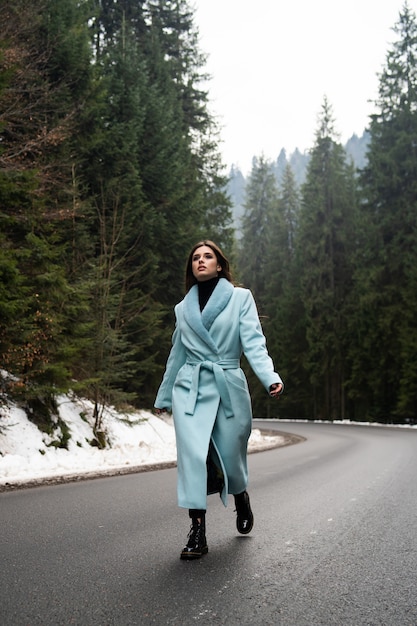 Garota morena sexy andando na estrada na mesa de montanhas de inverno. Glamourosa jovem vestindo elegante casaco longo azul. Moda, negócios e pessoas bonitas conceito ..