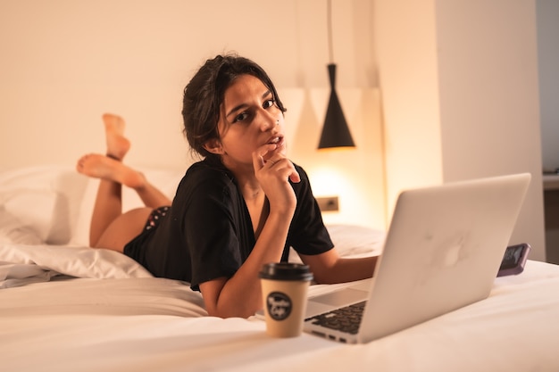 Garota morena sexy relaxada na cama com um computador e um café, lendo redes sociais