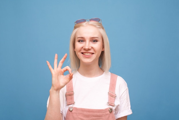 garota mostra um sinal de OK olha para a câmera e sorri, usa uma roupa casual fofa e óculos escuros, isolados no azul