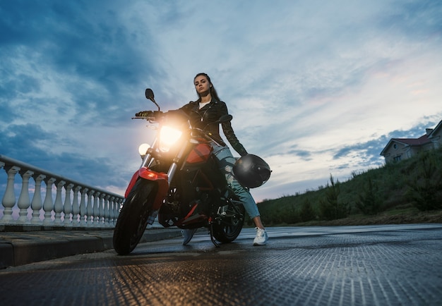 Garota motociclista em uma jaqueta de couro em uma motocicleta de cor preta e vermelha. bela paisagem no fundo