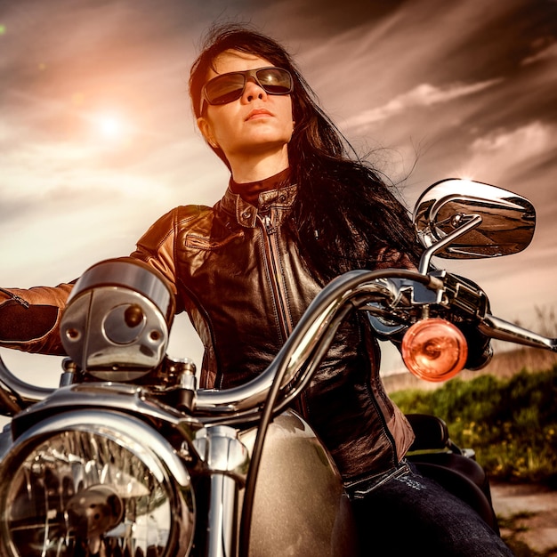 Garota motociclista em uma jaqueta de couro em uma motocicleta, olhando o pôr do sol.